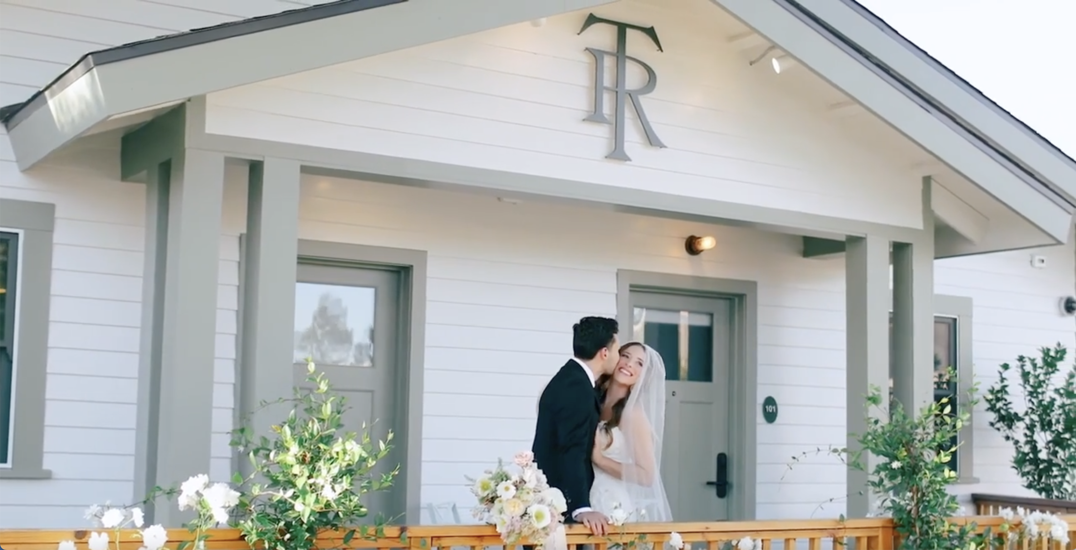 A white building with a gray trim and a couple in wedding attire standing in front of it.