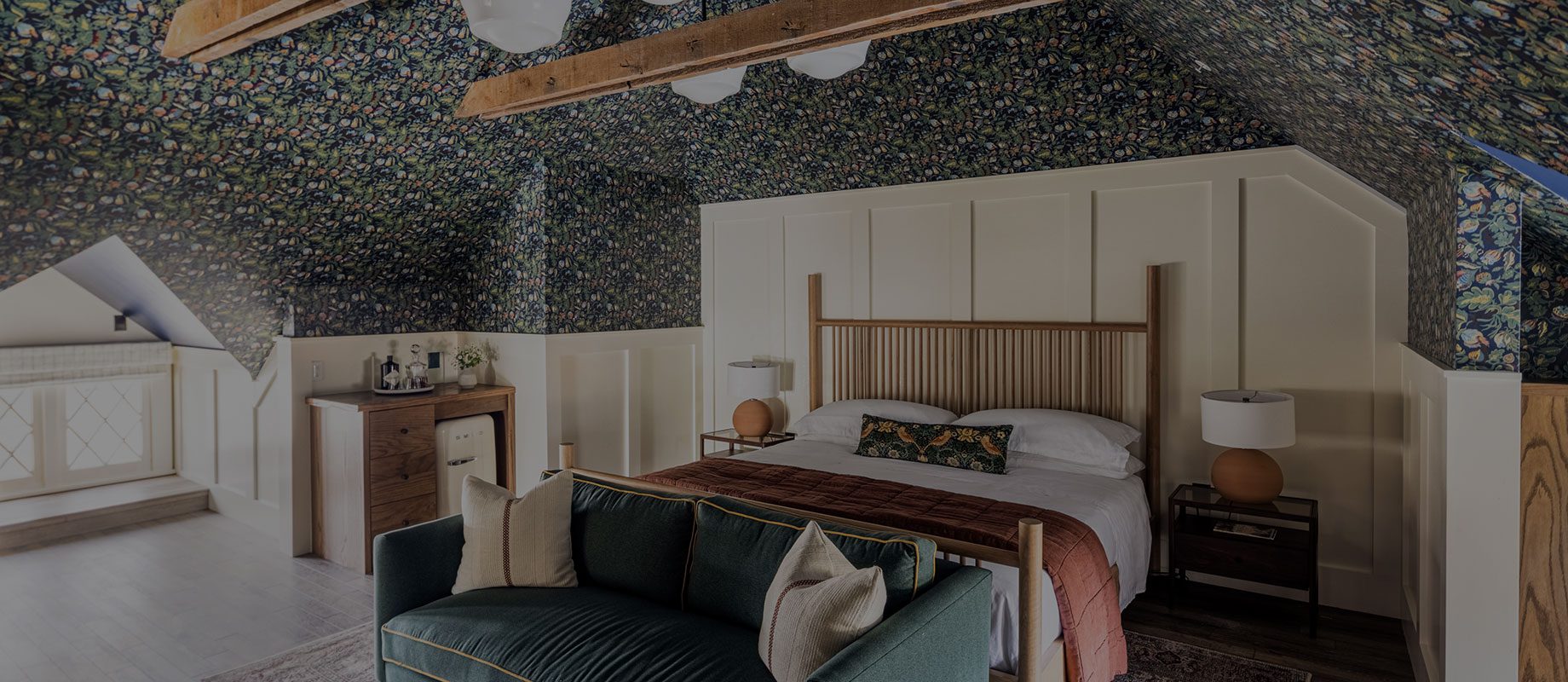 A cozy attic bedroom with floral wallpaper, exposed wooden beams, a wooden bed with white linens, a green sofa, bedside tables with lamps, and a small dresser near a window with built-in seating.